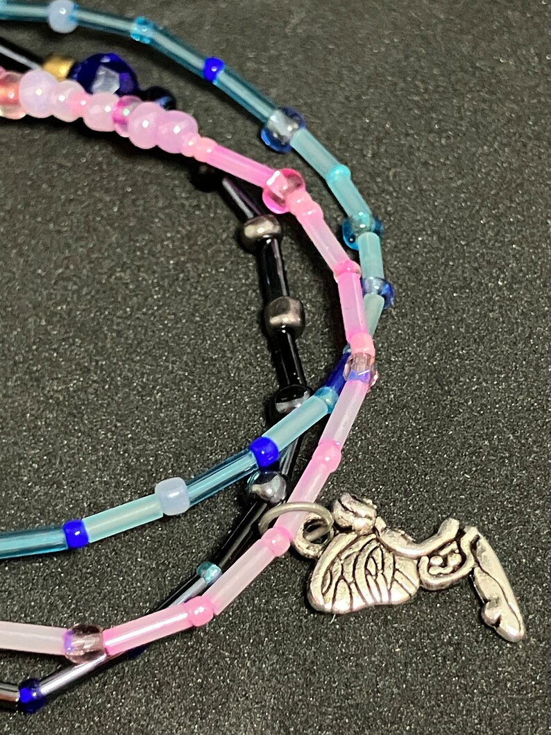 May include: A close-up of layered beaded necklaces in shades of pink, blue, and black. The necklaces feature small, colorful beads and a silver-toned charm shaped like a winged insect.