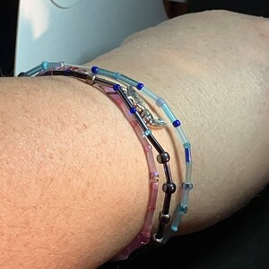 May include: Three beaded bracelets in shades of blue, purple, and black are wrapped around a wrist. The bracelets feature small, colorful beads strung on a thin wire, with a silver-colored charm on one. The bracelets are delicate and elegant.