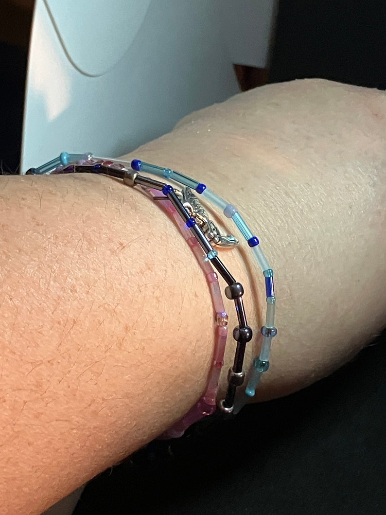 May include: Three beaded bracelets in shades of blue, purple, and black are wrapped around a wrist. The bracelets feature small, colorful beads strung on a thin wire, with a silver-colored charm on one. The bracelets are delicate and elegant.