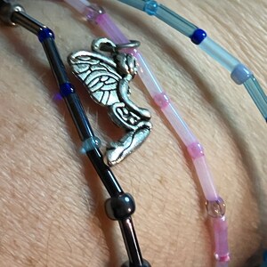 May include: Close-up of beaded bracelets in shades of blue, pink, and black. One bracelet features a silver fairy charm. The beads are small and cylindrical, with a mix of opaque and translucent colors. The bracelets are worn on a wrist.