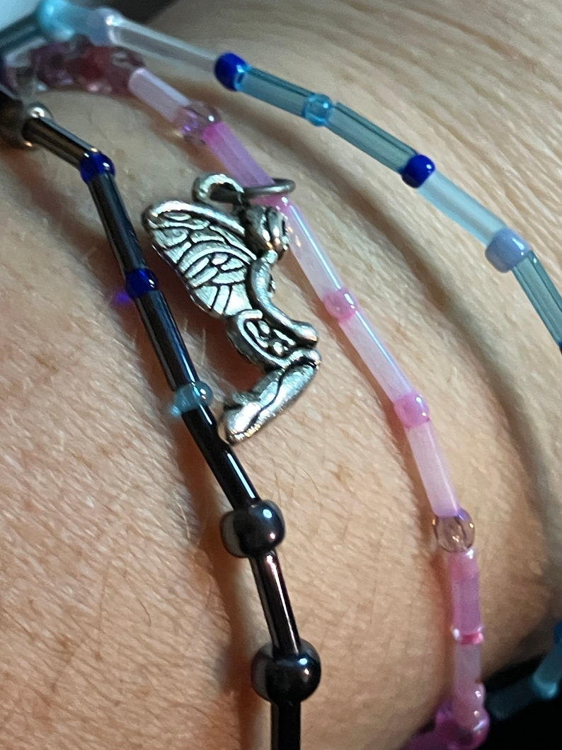 May include: Close-up of beaded bracelets in shades of blue, pink, and black. One bracelet features a silver fairy charm. The beads are small and cylindrical, with a mix of opaque and translucent colors. The bracelets are worn on a wrist.