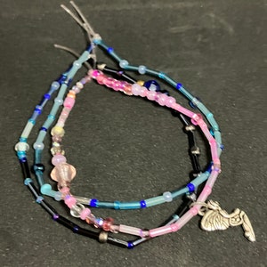 May include: Three beaded bracelets in shades of blue, pink, and black. The bracelets are strung with small beads and feature a silver charm. The bracelets are on a dark background.
