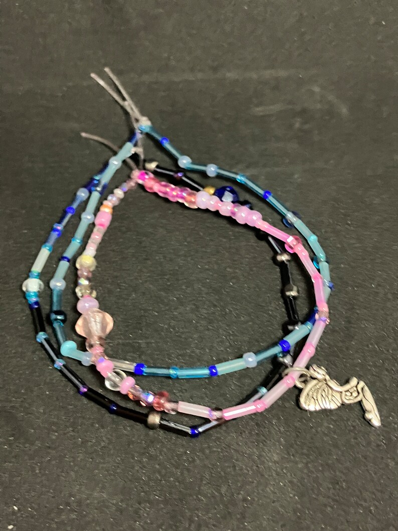 May include: Three beaded bracelets in shades of blue, pink, and black. The bracelets are strung with small beads and feature a silver charm. The bracelets are on a dark background.