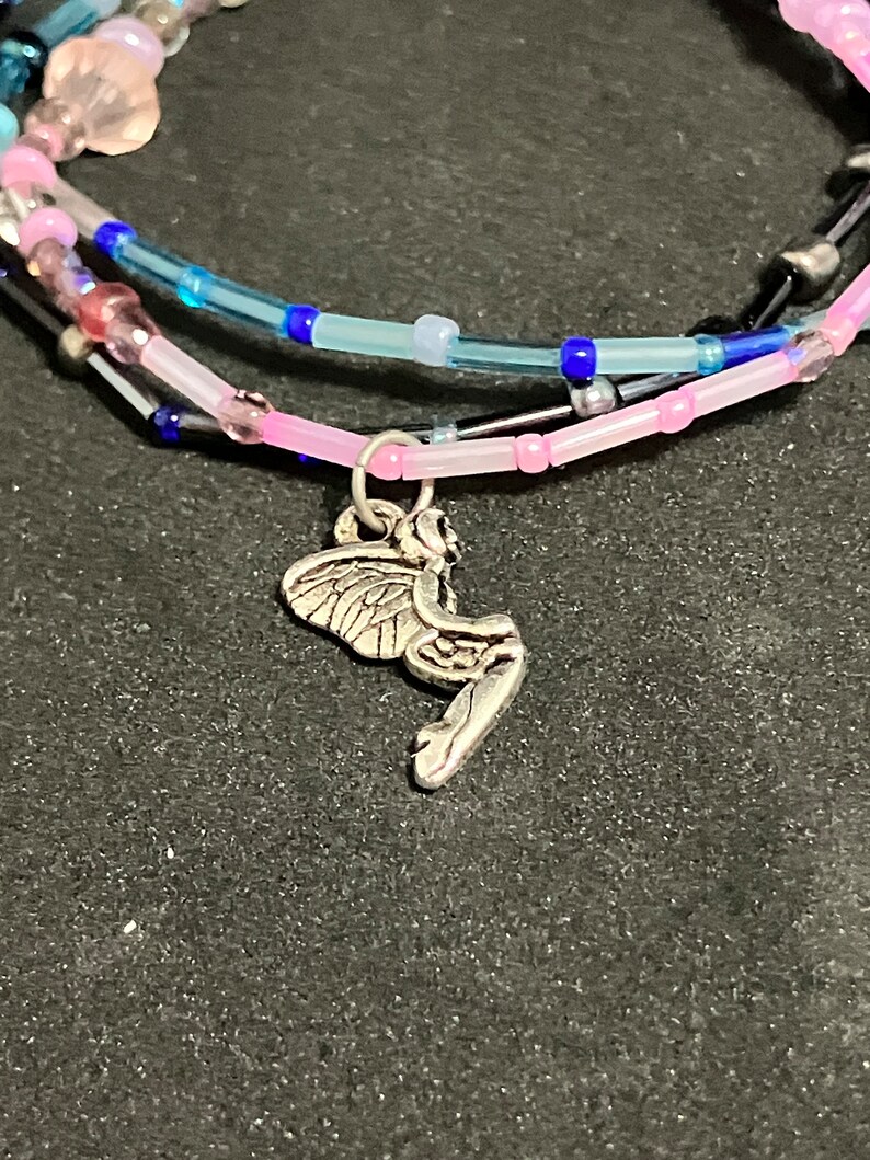 May include: A multi-strand bracelet featuring pink, blue, and clear beads. A silver fairy charm dangles from the bottom strand. The bracelet is on a dark surface.
