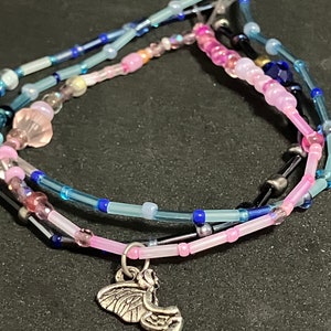 May include: A layered bracelet with pink, blue, and clear beads. A silver charm of a fairy hangs from the bottom layer. The beads are various sizes and shapes, with some iridescent and some opaque.