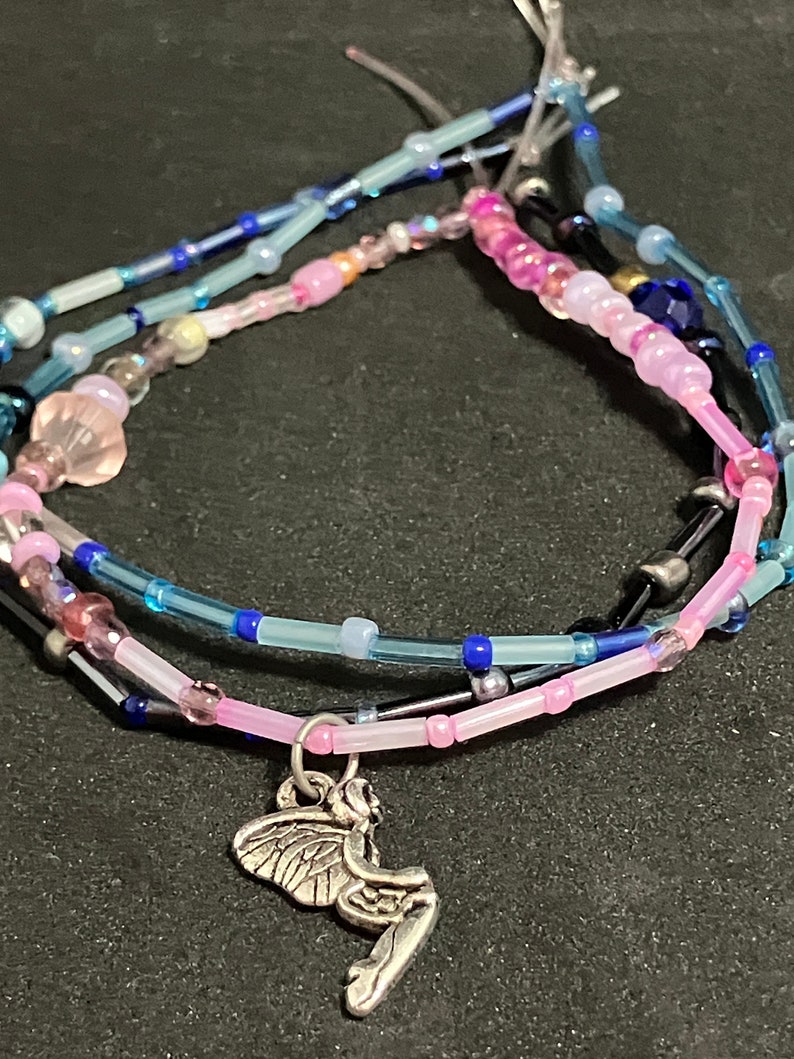 May include: A layered bracelet with pink, blue, and clear beads. A silver charm of a fairy hangs from the bottom layer. The beads are various sizes and shapes, with some iridescent and some opaque.
