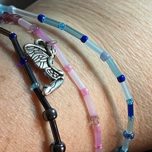 May include: Close-up of three beaded bracelets. One is black with dark beads and a silver angel charm. The other two are light pink and blue with small beads. The bracelets are worn together.