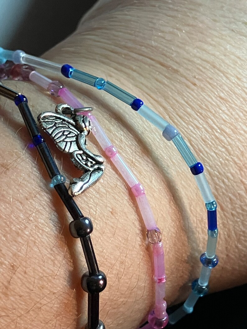 May include: Close-up of three beaded bracelets. One is black with dark beads and a silver angel charm. The other two are light pink and blue with small beads. The bracelets are worn together.