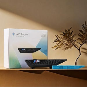 Könnte beinhalten: Ein BMW Natural Air Auto-Lufterfrischer-Starterset in einer weißen Schachtel mit geometrischen Farbakzenten. Das Set enthält einen schwarzen Halter mit M-Logo und einen blauen Nachfüllstab. Der Text auf der Schachtel lautet Natural Air.