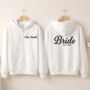 May include: Two white zip-up hoodies. One displays "Mrs. Smith" in black script on the front. The other has "Bride" and the date "29.5.2026" in black script on the back. Both have a hood and front pockets.