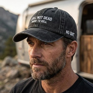 以下が含まれることがあります： 前面に「STILL NOT DEAD HARD TO KILL」、側面に「NO QUIT」と書かれた濃いグレーの野球帽。帽子は使い古されたような、少し色あせた外観をしています。