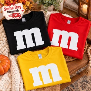 Puede incluir: Tres camisetas de cuello redondo en negro, rojo y amarillo, cada una con una letra m minúscula grande de color blanco impresa en la parte delantera. Las camisetas están dobladas y dispuestas juntas, mostrando los colores sólidos y la tipografía blanca centrada.