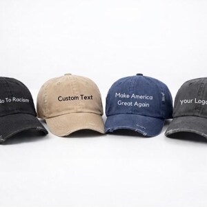 May include: Four distressed baseball caps in black, tan, and navy blue. Each cap has text: "Say No To Racism", "Custom Text", "Make America Great Again", and "your Logo Here". The caps have a worn, vintage look.