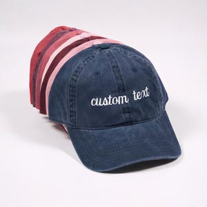 May include: A stack of baseball caps in various shades of red and blue. The frontmost cap is navy blue with the embroidered text "custom text" in white. The other caps are shades of red and pink.