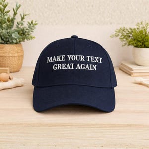 May include: A navy blue baseball cap with the text "MAKE YOUR TEXT GREAT AGAIN" embroidered in white. The cap has a curved brim and a button on top. The hat is sitting on a wooden surface.