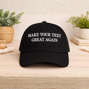 May include: Black baseball cap with the text "MAKE YOUR TEXT GREAT AGAIN" in white. The cap has a curved brim and a button on top. The hat is sitting on a light-colored wooden surface.