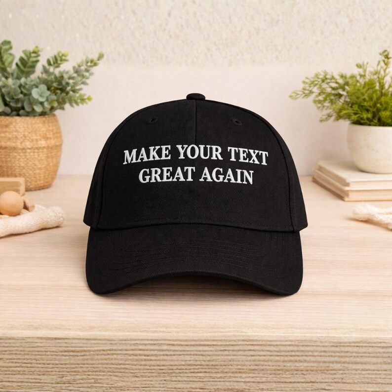 May include: Black baseball cap with the text "MAKE YOUR TEXT GREAT AGAIN" in white. The cap has a curved brim and a button on top. The hat is sitting on a light-colored wooden surface.