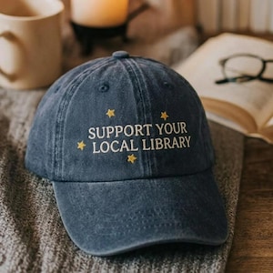 May include: A navy blue baseball cap with the embroidered text "SUPPORT YOUR LOCAL LIBRARY" in white and yellow star accents. The cap is made of a soft, worn fabric and has a curved brim. A stylish accessory for book lovers.