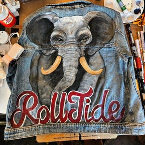 May include: A denim jacket with a hand-painted elephant design in shades of gray, with yellow tusks. The words "Roll Tide" are painted in red script across the bottom. The jacket is a light blue wash.
