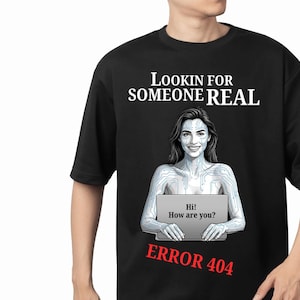 以下が含まれることがあります： 黒いTシャツ。肌に回路基板の模様がある人物が、「Hi! How are you?」と表示されたノートパソコンを持っているグラフィックが描かれています。上部には「LOOKIN FOR SOMEONE REAL」、下部には赤い文字で「ERROR 404」とプリントされています。