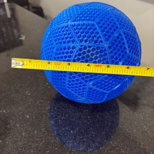 May include: A bright blue spherical object with a geometric honeycomb lattice pattern. A yellow measuring tape is positioned across the center of the sphere, indicating a diameter of approximately 6.5 inches from edge to edge.