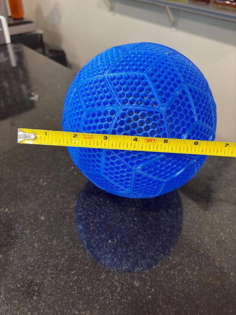 May include: A bright blue spherical object with a geometric honeycomb lattice pattern. A yellow measuring tape is positioned across the center of the sphere, indicating a diameter of approximately 6.5 inches from edge to edge.