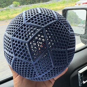 May include: A dark blue spherical object featuring a complex geometric lattice structure. The surface is composed of repeating hexagonal patterns organized into larger pentagonal and hexagonal panels, creating a hollow, open-work design.