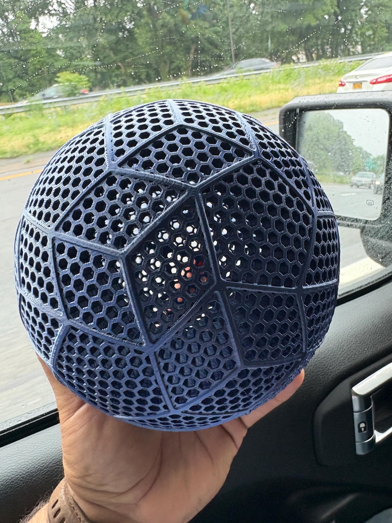 May include: A dark blue spherical object featuring a complex geometric lattice structure. The surface is composed of repeating hexagonal patterns organized into larger pentagonal and hexagonal panels, creating a hollow, open-work design.