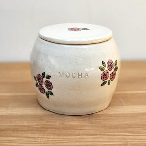May include: White ceramic jar with a lid, decorated with pink roses and green leaves. The jar has the word "MOCHA" written on the front.