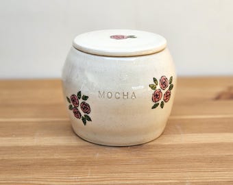May include: White ceramic jar with a lid, decorated with pink roses and green leaves. The jar has the word "MOCHA" written on the front.