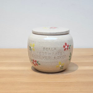 May include: A white ceramic urn with a lid, decorated with colorful flowers and the inscription "Bella 2008-2021 Forever Loved".