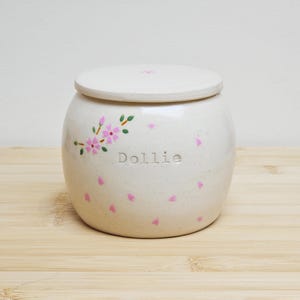 May include: A cream-colored ceramic jar with a matching lid. The jar is decorated with pink floral accents and pink petal shapes. The name "Dollie" is inscribed on the front. The jar is sitting on a light wood surface.