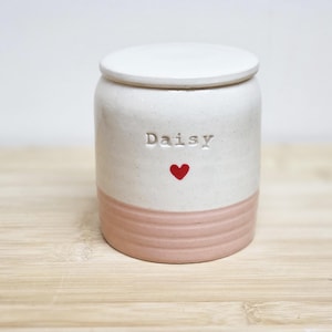 以下が含まれることがあります： 蓋付きの陶器製のジャー。上部はオフホワイトで、「Daisy」という名前と赤いハートが付いています。下部は、水平の溝がある薄いピンク色です。装飾的な収納容器。