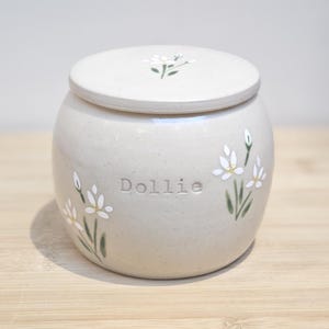Puede incluir: Jarra de cerámica beige claro con tapa, adornada con flores de margarita blancas y tallos verdes. El nombre "Dollie" está grabado en relieve en la parte delantera. Diseño sencillo y elegante.
