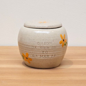 Puede incluir: Un tarro de cerámica blanco con tapa, decorado con flores amarillas y el texto "Onion 04 FEB 08 TO 04 MAR 21".