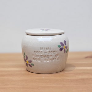 May include: White ceramic urn with a lid, decorated with purple and yellow flowers. The urn has the inscription "Mimi 2006 - 2021 Forever in Our Hearts".