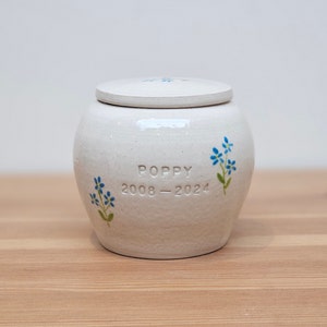 May include: White ceramic pet urn with a lid, featuring blue forget-me-not flowers and the inscription "Poppy 2008-2024".
