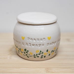 Puede incluir: Un pequeño tarro de cerámica blanquecina con tapa. El tarro está decorado con flores amarillas y hojas verdes alrededor de la base. Las palabras "Peanut Forever & Always Baby" están grabadas en la parte delantera, con corazones amarillos.