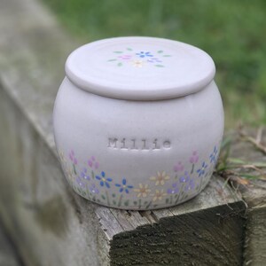 May include: A pale grey ceramic jar with a lid, adorned with hand-painted flowers in pastel hues. The name "Millie" is embossed on the jar. The jar rests on a weathered wooden surface.