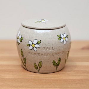 May include: A small, white ceramic urn with a lid, decorated with white daisies and green leaves. The urn has the words "Ball Forever Loved" engraved on the side.