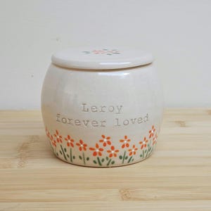 Puede incluir: Un tarro de cerámica color crema con tapa, adornado con motivos florales naranjas y verdes. El tarro lleva la inscripción "Leroy forever loved". La tapa también presenta pequeños diseños florales.