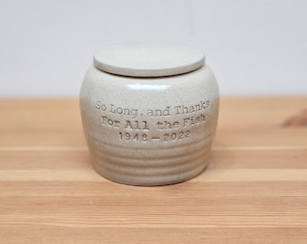May include: A light beige ceramic jar with a matching lid, featuring a speckled finish and horizontal ridges at the base. The text "So Long, and Thanks For All the Fish" and the dates "1948-2022" are engraved into the front of the vessel.