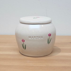 May include: White ceramic jar with a lid, featuring a floral design with pink tulips and green stems. The jar is inscribed with the name "MARTHA".