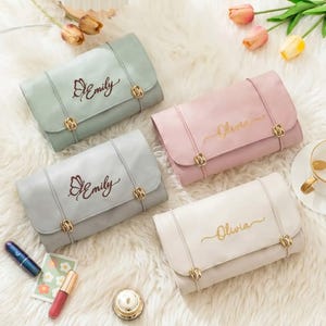 Puede incluir: Cuatro carteras de mano de colores pastel con herrajes dorados. Dos carteras tienen el nombre "Emily" con un diseño de mariposa, y dos tienen el nombre "Olivia". Las carteras están dispuestas sobre una superficie blanca y esponjosa.