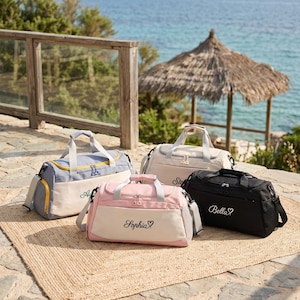 Könnte beinhalten: Vier Reisetaschen in Blau, Beige, Rosa und Schwarz, jeweils mit einer Vordertasche, zwei Tragegriffen und einem abnehmbaren Schultergurt. Die rosa Tasche zeigt den Namen Sophia mit einem Herz, die schwarze Tasche den Namen Bella mit einem Herz.