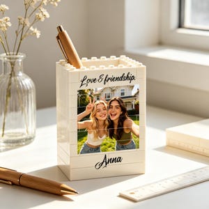 Könnte beinhalten: Ein weißer, rechteckiger Stiftehalter aus ineinandergreifenden Bausteinen. Die Vorderseite zeigt ein aufgedrucktes Foto von zwei lächelnden Personen, den Text "Love & friendship." über dem Foto und den Namen "Anna" darunter gedruckt.