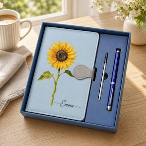 Könnte beinhalten: Ein hellblaues Notizbuch mit einer gelben Sonnenblumenillustration und dem Namen Emma in schwarzer Schreibschrift. Das Notizbuch hat einen grauen Magnetverschluss und liegt in einer blauen Geschenkbox neben einem blauen Stift und einer Mine.