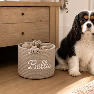 May include: A round, beige woven rope storage basket featuring two knotted handles and the name Bella embroidered in white script on the front. The basket contains several small, textured toys and sits next to a black, white, and tan dog.