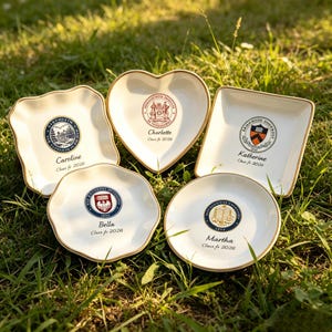 May include: Five white ceramic dishes with gold rims, each with a different shape and design. The dishes feature university crests and names like "Caroline" and "Katherine", with "Class of 2026" below. They are arranged on green grass.