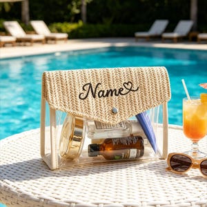 May include: A clear cosmetic bag with a woven beige top flap, featuring the word "Name" in black script with a heart. The bag contains various beauty products. A refreshing orange drink and sunglasses are nearby.
